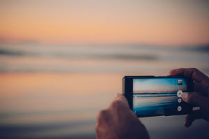 Hands holding a phone and taking a picture of a sunset over a beach