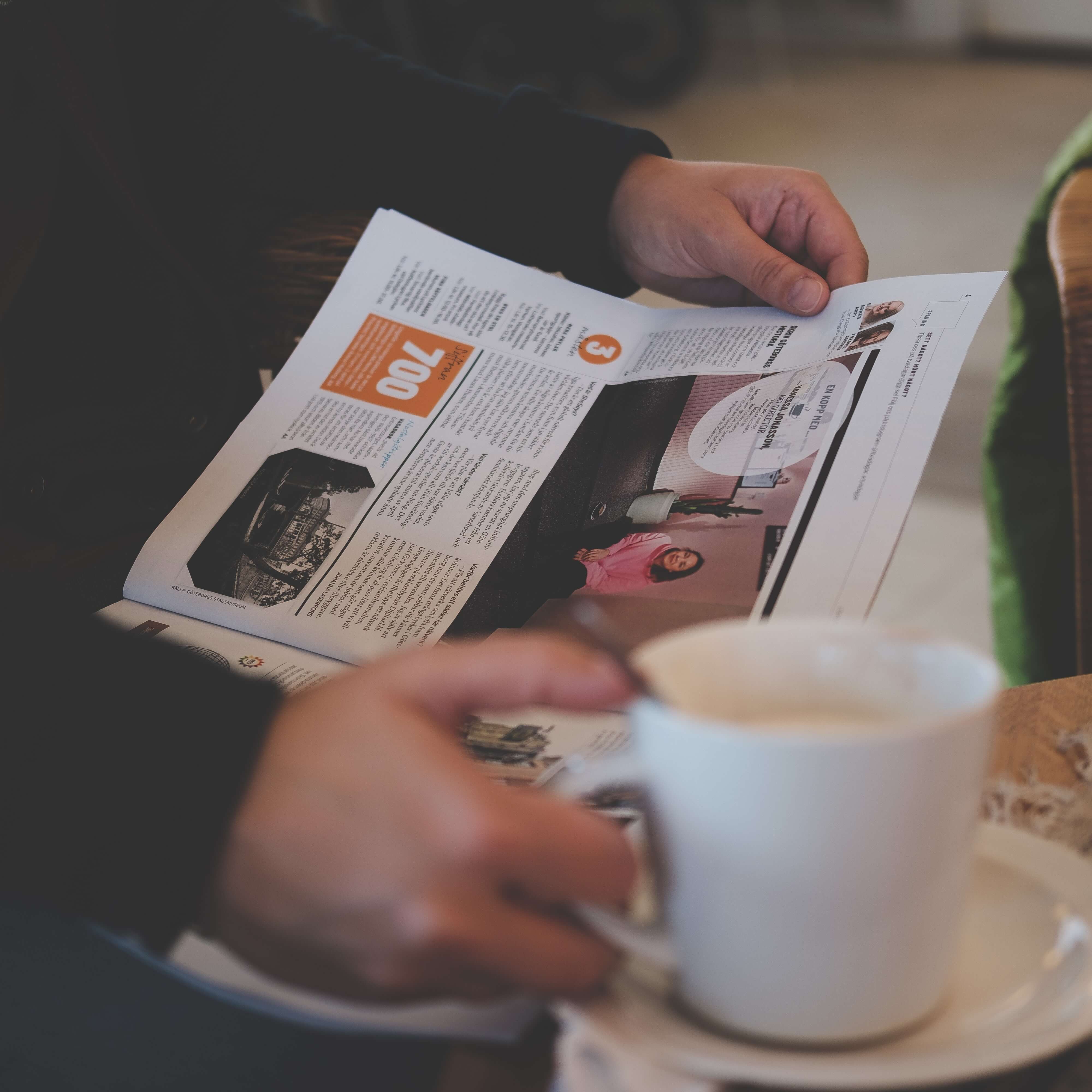 A person reads the news paper whilst holding a cup.