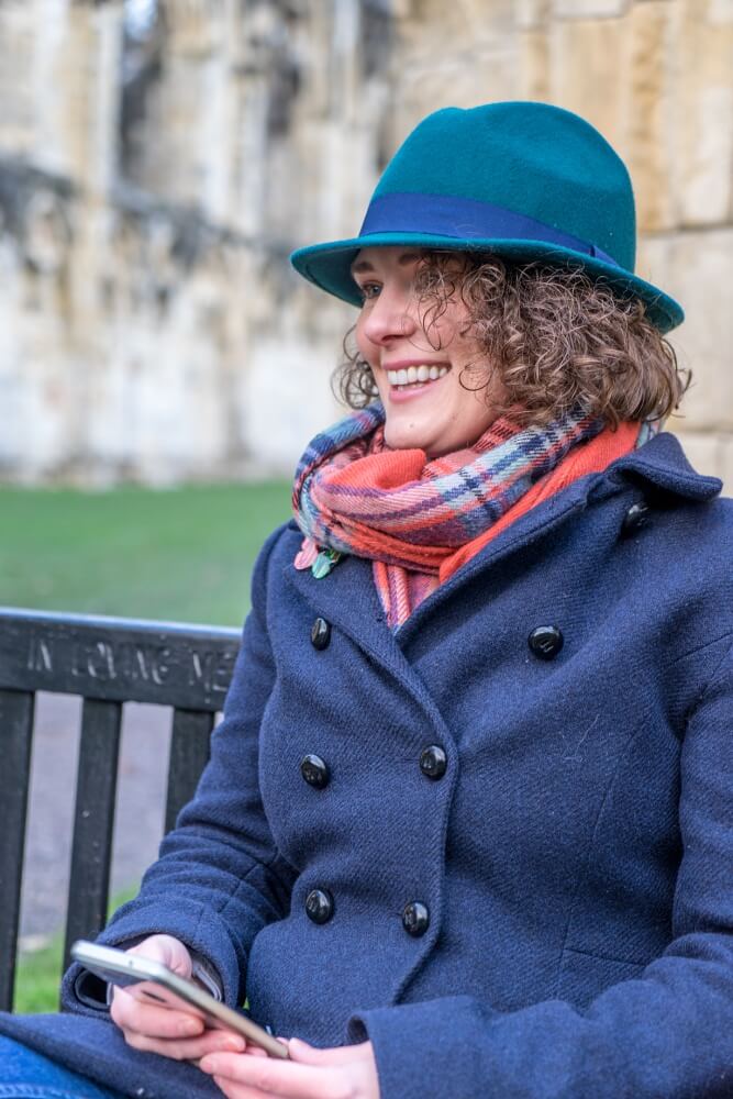A photo of Amy Jeffs sitting on a bench in a blue coat & hat smiling.
